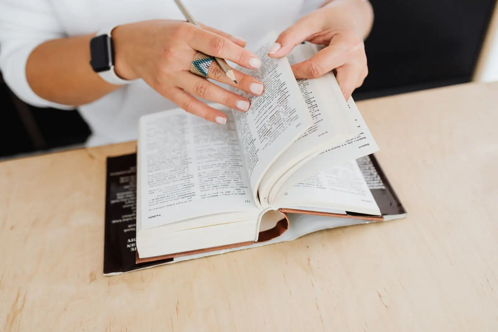 Mains feuilletant un dictionnaire ouvert, un crayon à la main, sur un bureau en bois