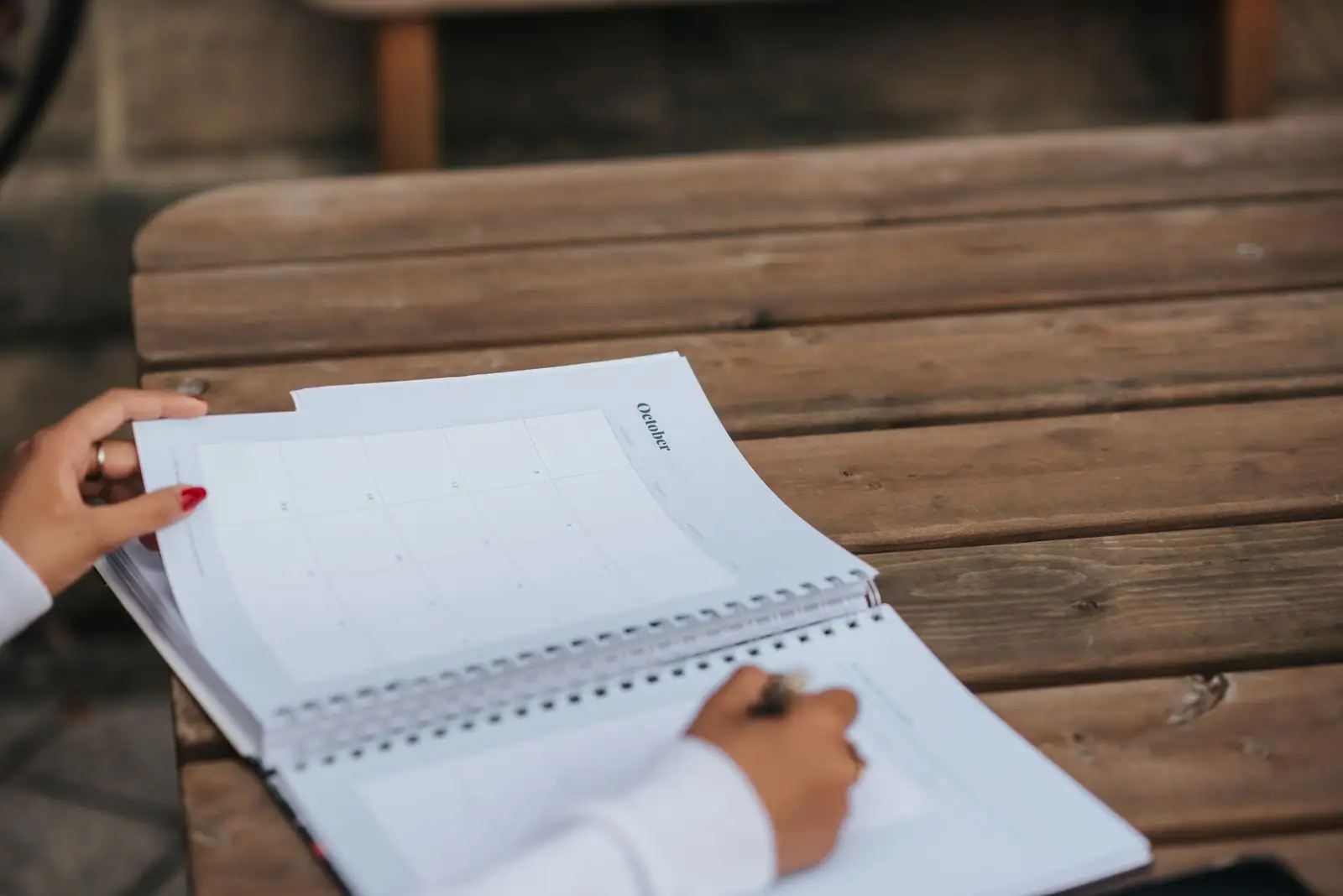 Mains feuilletant un calendrier mensuel et un carnet à spirale, un stylo à la main, sur une table en bois.