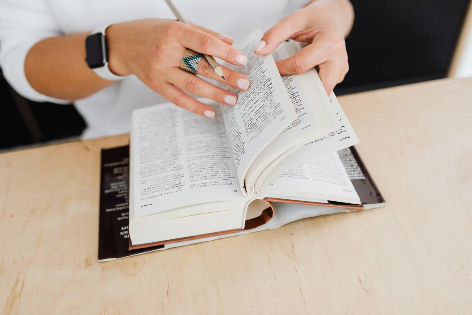 Mains feuilletant un dictionnaire épais sur un bureau, à la recherche du mot juste, crayon en main