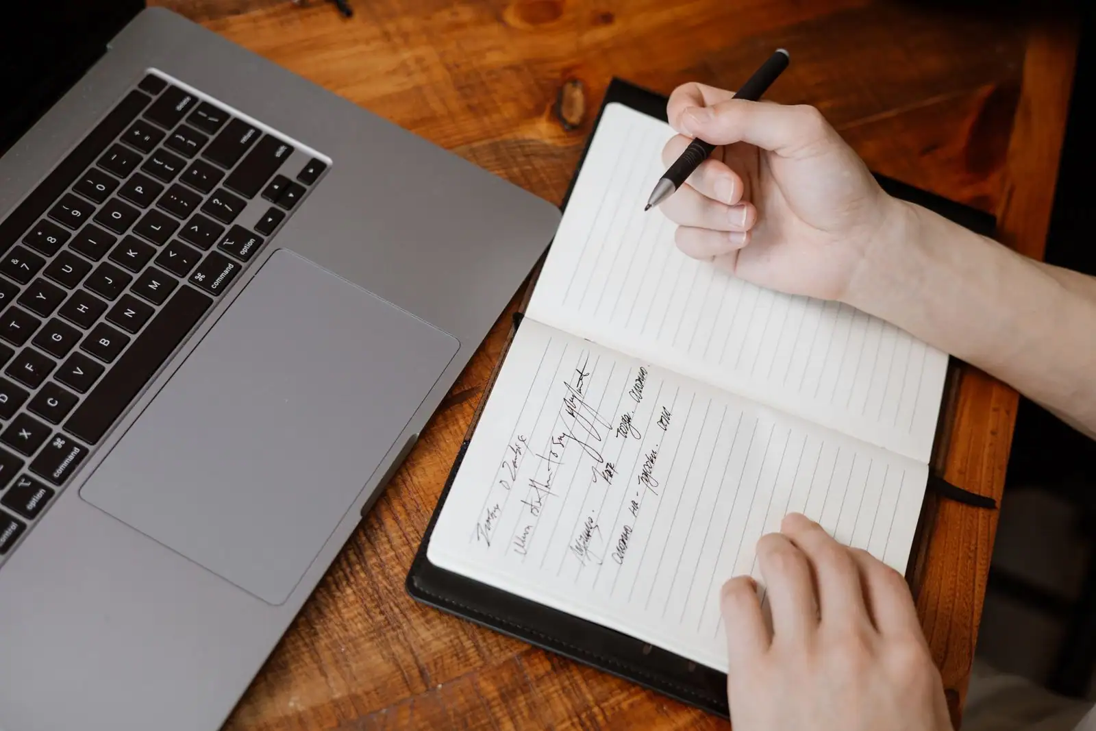 Mains rédigeant des notes dans un carnet, un ordinateur portable posé à côté sur un bureau en bois.