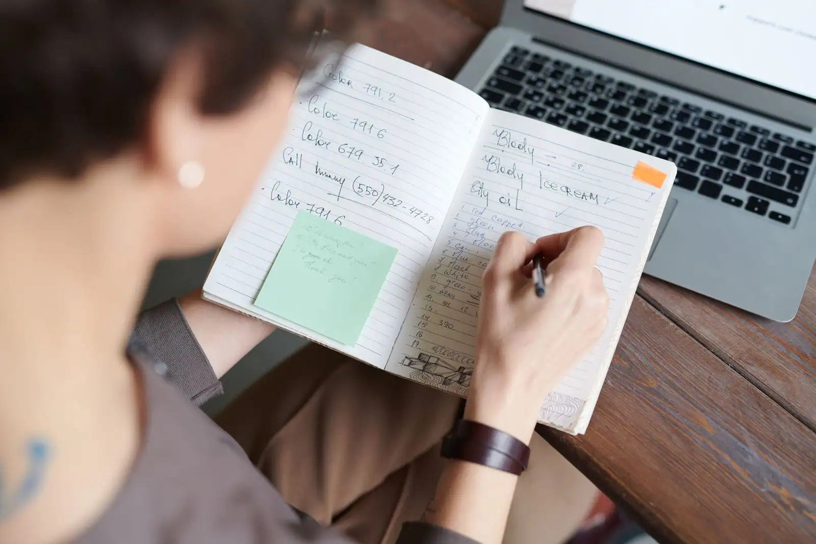 Personne prenant des notes dans un carnet, un ordinateur portable posé à côté sur un bureau