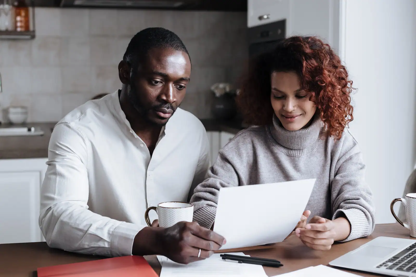 Couple attablé dans une cuisine, examinant ensemble un document papier à côté d'un ordinateur portable.