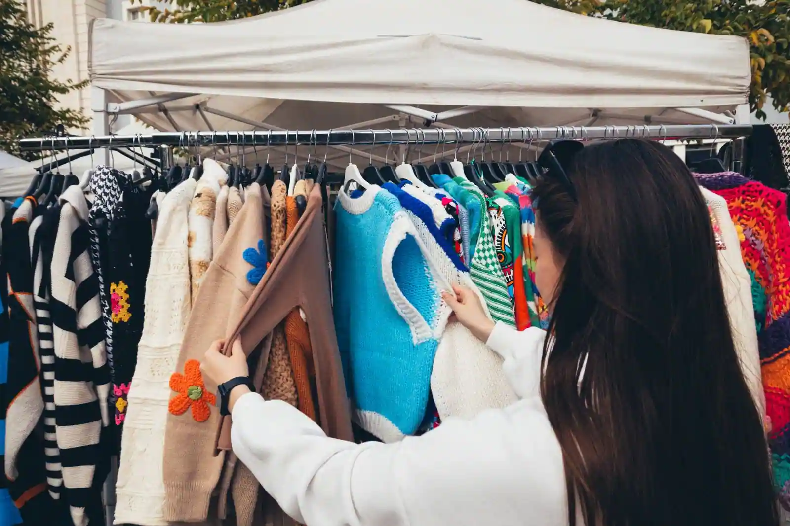 Une femme parcourt un portant de pulls et gilets d'occasion sur un stand de marché abrité par une tonnelle