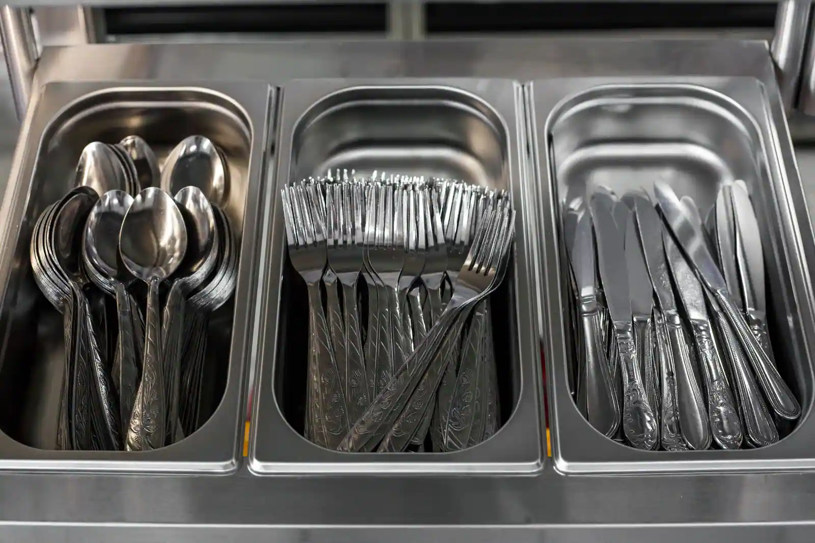 Bacs en inox alignés sur la ligne de self d'une cantine, garnis de cuillères, fourchettes et couteaux.