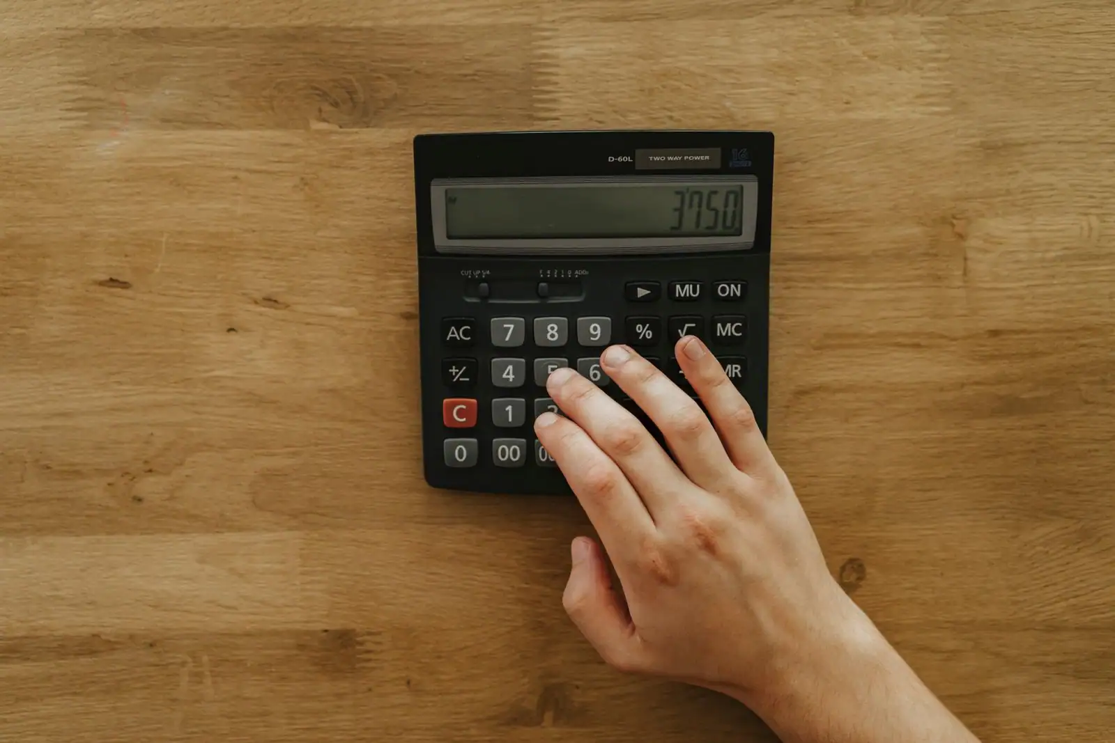Une main compose un calcul sur une calculatrice de bureau noire posée sur un plan de travail en bois.