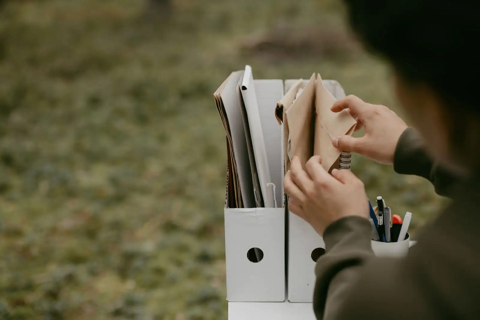 Des mains classent des documents dans deux porte-revues cartonnés, sur un fond neutre et flouté.