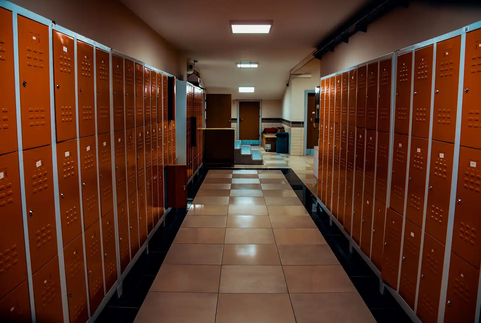 Vestiaire de club désert, deux rangées de casiers orange de part et d'autre d'une allée carrelée.