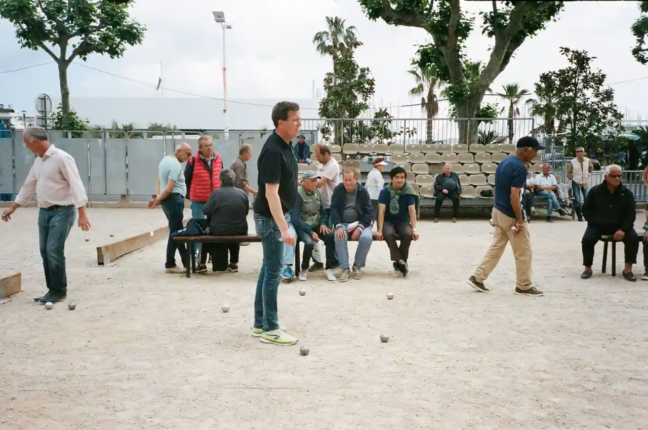 Joueurs de pétanque sur un boulodrome en terre, boules métalliques au sol et spectateurs assis sur des bancs.