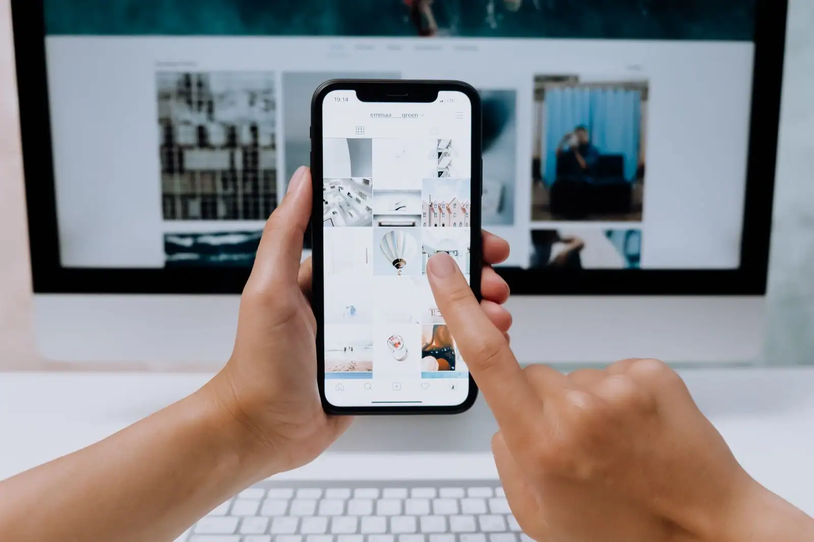 Une main tient un smartphone affichant un profil de réseau social devant un écran d'ordinateur ouvert sur un site.