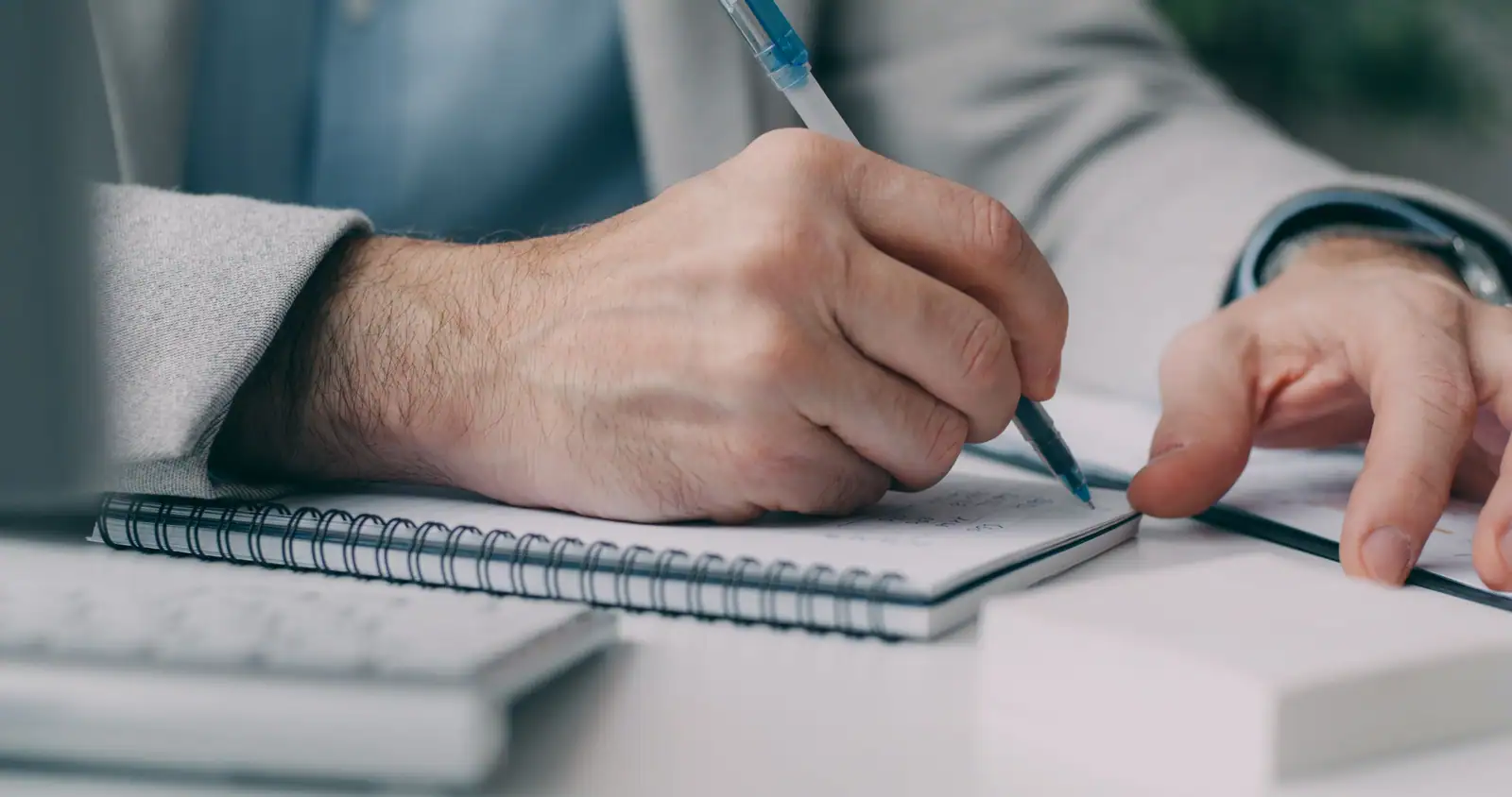 Une personne en veste prend des notes dans un carnet à spirale posé près d'un clavier d'ordinateur.
