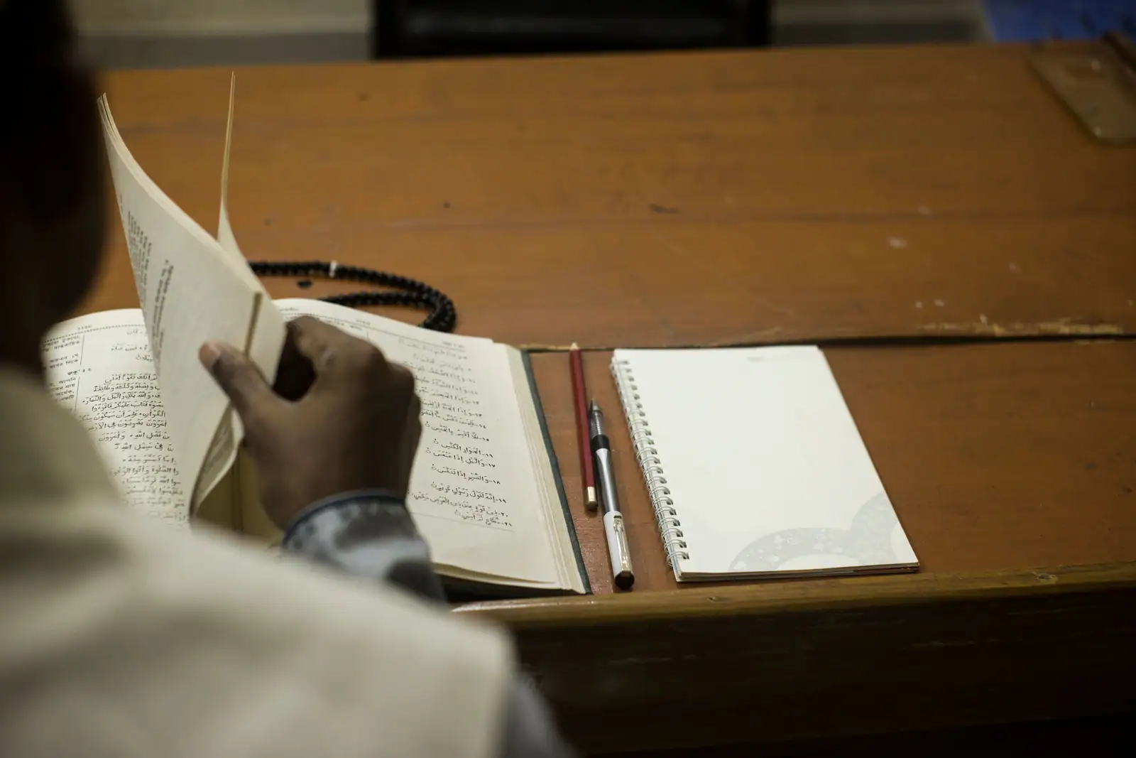 Une personne tourne les pages d'un ouvrage religieux en arabe, près d'un chapelet et d'un carnet vierge.